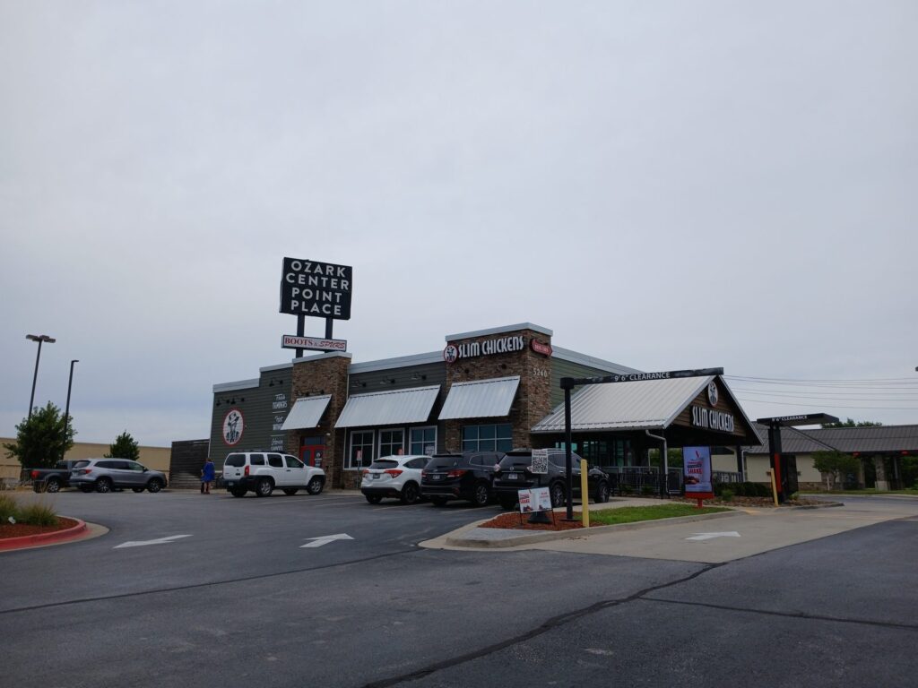 Exterior of Slim Chickens restaurant at Ozark Center Point Place with a row of parked cars in front on a cloudy day